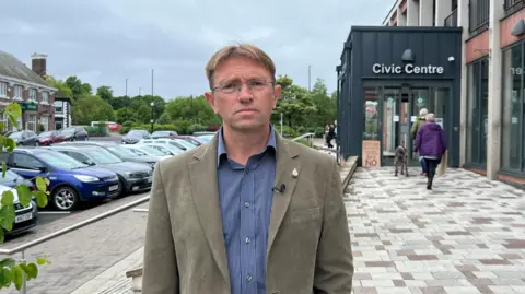 Geoff Moss is wearing glasses, a beige jacket and a blue and white striped shirt. He is standing in front of Carlisle Civic Centre. He does not look happy.