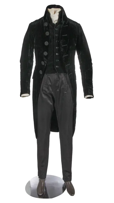 Cosprop and Kerry Taylor Auctions An early 19th century style suit comprising a cutaway black velvet tailcoat, large moulded glass jet-effect buttons, a closely fitting un-patterned black velvet waistcoat, a pair of black satinised wool trousers with elasticated stirrup bands to hems and a cream cotton shirt with matching stock.