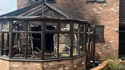 Emma Parker The burnt out remains of the conservatory. The window frames remain, but there is very little glass. The metal frames of chairs can be seen inside. There is rubble on the floor and the brickwork behind the conservatory is singed. 