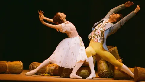Getty Images Marianela Núñez as Lise in a white dress and Vadim Muntagirov as Colas in a floral waistcoat, blue jacket and yellow tights - they are in rehearsal for a Royal Ballet performance of Frederick Ashton's La Fille Mal Gardée at the Royal Opera House in October 2025.