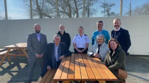 (L-R) John De Carteret (prison governor), deputy Rob Prow, prison officer Birkett, prison officer Mel Fletcher, Deputy Susan Aldwell, Dave Le Feuvre (head of education, training and regimes), Michelle Watson Bunn (Ogier Law Firm) and Christopher Jones (Ogier Law Firm). The group are sat on one of the wooden benches made at the prison on a sunny day.