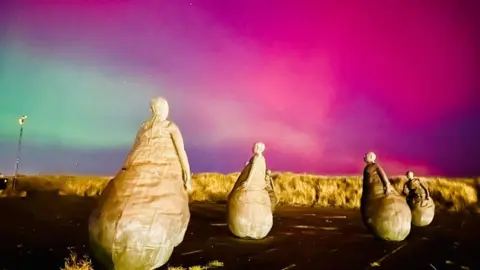 Aaron Aldus The Weebles - several bronze art pieces - are lit up by the beach in South Shields. The background sky is lit up green, purple and bright pink by the Northern Lights.