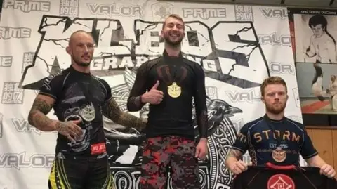 PA Media Will Stone pictured at a MMA competition. He stands in between two men and poses with a medal around his neck.