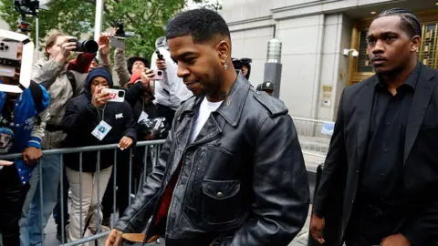 Rapper Kid Cudi in a leather jacket walking outside of a building as people with phones and cameras photograph him in the background. 