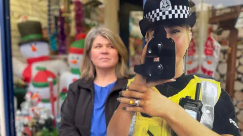 A cardboard cut out of a female police officer pointing a speed camera at the picture. She is wearing a black and white police hat and yellow hi-vis jacket. Anne Ness is in the background and they are in the door way of her shop.