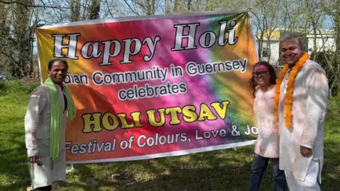 Indian Community in Guernsey Three people, two men and one woman, standing in white clothing next to a brightly coloured sign which says 'Happy Holi, Indian Community in Guernsey celebrates, Holi Ustav, festival of colours, love and joy. 
