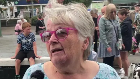 Wendy Chadwick wearing pink-rimmed glasses. She is standing in front of a crowd of people in Dawlish town centre