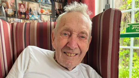 Darren Rozier/BBC An elderly man with short white hair and white T-shirt sits in a red stripey armchair. Behind him is a window looking out on to green trees and bushes. 