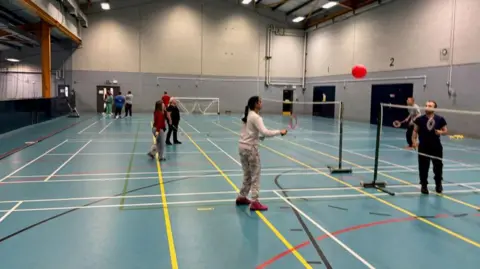 Bedford Borough Council People playing badminton in a large hall inside a leisure centre. The floor is blue with multi-coloured lines.