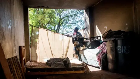 BBC/SHIRAAZ MOHAMED An unidentified woman stands in a room containing a mattress and other minimal essentials in the derelict San Jose building in Johannesburg, South Africa