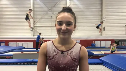 Jonathan Park/BBC Girl with dark hair tried up, wearing a gym vest. Trampolinists are visible in the background