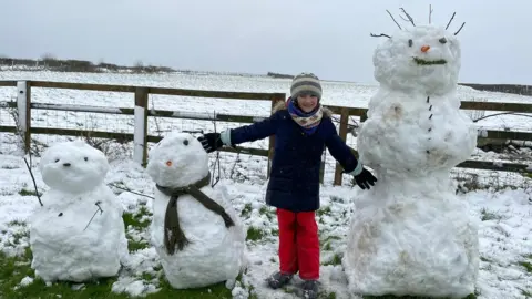 Andrew Carbis A child stands in the middle of a row of three snowmen with their arms held out to the side