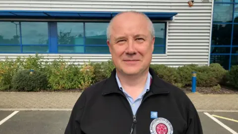 Smiling man with very short light hair stands outside an office building wearing a blue zip-up jacket