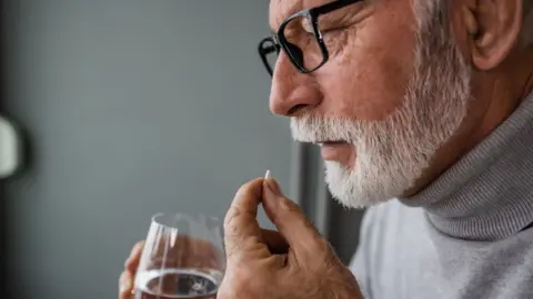 A man with a white beard holds a glass of water while taking a pill