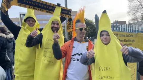 BBC Four people, three dressed in banana costumes hold up their thumbs and smile at the camera outside a stadium