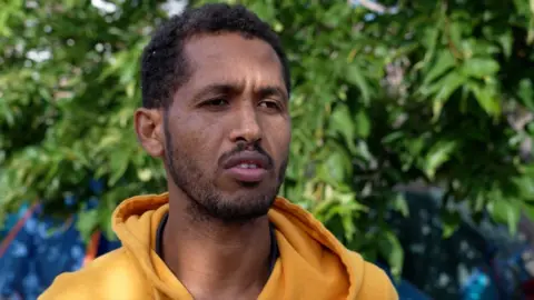 A man in a light orange hoodie talks during an interview