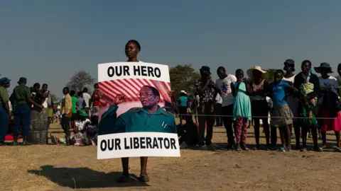Getty Images A young woman holds a placard reading 'Our hero, our liberator' villager queue to view the coffin of late former Zimbabwean president Robert Mugabe lying in state at Murombedzi Growth Point, about 107 km northwest of Harare, Zimbabwe, on September 16, 2019, as people have been accorded the opportunity to view Mugabe's body a week after his death. - The remains of former Zimbabwe president Robert Mugabe were taken to his village for a wake on September 16, a family member said, as his final burial is prepared in about a month. Mugabe died a week ago aged 95 in Singapore, nearly two years after he was ousted in a 2017 coup that ended nearly four decades of increasingly autocratic rule. After a state funeral on September 14 in the capital Harare attended by African leaders, his body went to his rural village of Kutama, 90 kilometres (55 miles) to the west, to allow villagers to pay tribute and bid farewell.