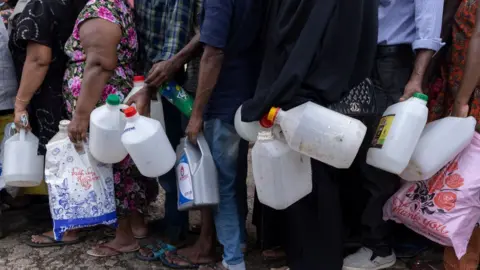 Getty Images People hold empty bottles while they wait in a queue for several hours to buy fuel amid the economic crisis at a gas station on May 18, 2022 in Colombo, Sri Lanka.