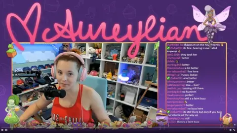 Aureylian/Twitch A screenshot of a Twitch stream. In the middle is the video stream featuring Aureylian, a young woman wearing a headset in front of a microphone. In the background are various gaming consoles and controllers, as well as figurines and models. The screenshot also shows a chat screen filled with various comments from her fans.