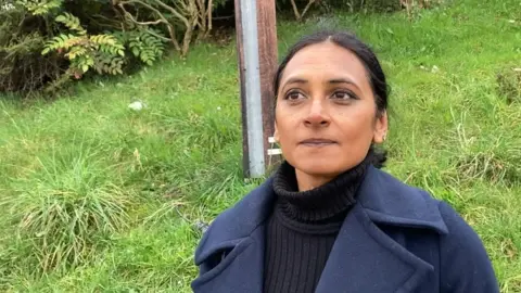 Meena Powell in a blue coat standing waiting for the bus