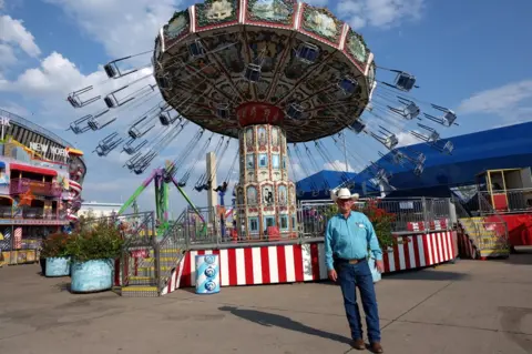 BBC A man in a cowboy hat stands in front of a fair ride