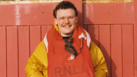 Geoff Cowan A man with a moustache and glasses wearing a yellow jacket and red life vest smiles at the camera