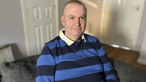 BBC/Munaza Rafiq Picture of Adam sitting on a sofa in his living room. He's is wearing a striped blue and black polo shirt