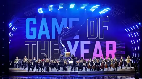 Getty Images An orchestra performs on a large stage with "Game of the Year" projected on to a screen at the rear in colourful block capital letters. An image of the awards statue - a winged figure leaning backwards as if about to take flight - is superimposed on top. There are between 30 and 40 members of the orchestra, most of them seated and playing a range of stringed and woodwind instruments. To the far right, an electric guitarist stands facing the crowd, and a large cylindrical Japanese Taiko drum is on a plinth in the centre of the group.