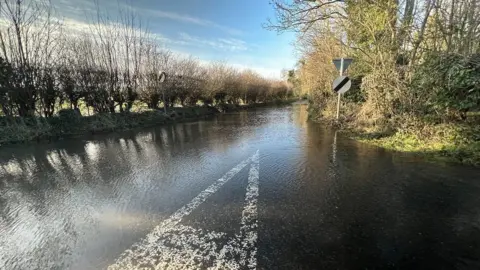 BBC A flooded road in the Wiltshire countryside, hedges down the side with light reflecting off the water and a blue sky