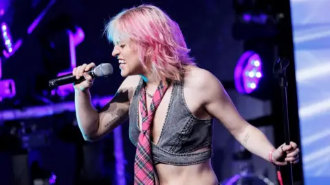 Getty Images Beth McCarthy performing on stage. Beth is a 26-year-old white woman with blonde hair dyed pink. She wears a grey denim crop top with a pink tartan tie around her neck. She holds a microphone to her face with her right hand, revealing a black line tattoo on her inner upper arm. Beth's eyes are closed as she sings, holding the microphone stand with her left hand. The staging behind her is lit purple