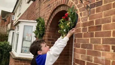 Martin Heath/BBC Boy dressed in blue and white reaches up to hang a holly wreath beside a front door