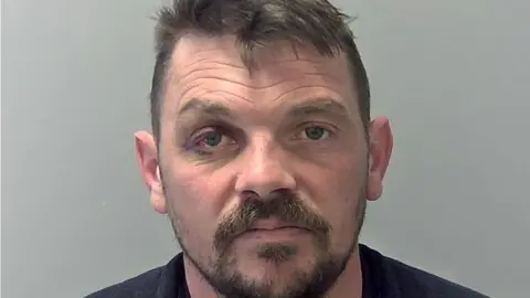 Suffolk Police Mugshot of David Perry. He is white with dark hair and a bloodshot right eye, He also has a dark moustache and short beard.