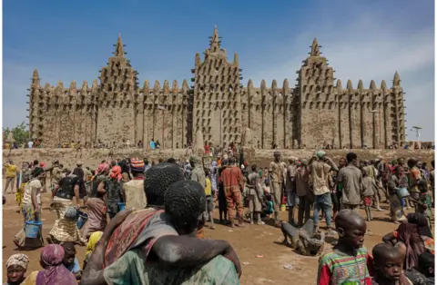 Ousmane Makaveli/AFP People staring at the Great Mosque of Djenne - 4 June. It is a large brown building which stands against a backdrop of blue skies.