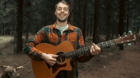 Nuart Photography Sam Evans holding a guitar in a forest