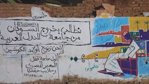 BBC A mural with the words: "We demand that Sudan leaves the Arab League. We are black people, the sons of Kushites and the return of the Hala'ib Triangle" - Khartoum, Sudan