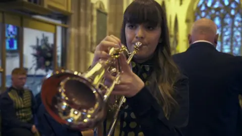 BBC Teenage girl with long dark hair plays flugelhorn solo with band and conductor behind her