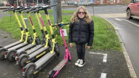 BBC Ella standing next to scooters