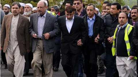 PA Media Dr Abdul Shakoor walks immediately behind the funeral cortege with a large group of people, as it arrives at Parndon Wood Cemetery, in Harlow, for his family's burial following their funeral service at Harlow Islamic Centre