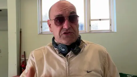 Still of Michael Monaghan, wearing sunglasses and headphones around his neck, at the foodbank.