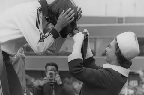 Getty Images Queen Elizabeth gets the ribbon entangled in the hair of Debbie Brill of Canada, as she presents her with the gold medal for the high jump at Meadowbank Stadium, during the Commonwealth Games in 1970