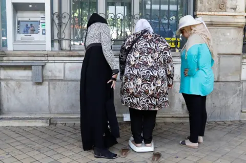 Reuters A woman on a weighing scale outside a bank observed by two other women in Tunis, Tunisia - Thursday 30 August 2018
