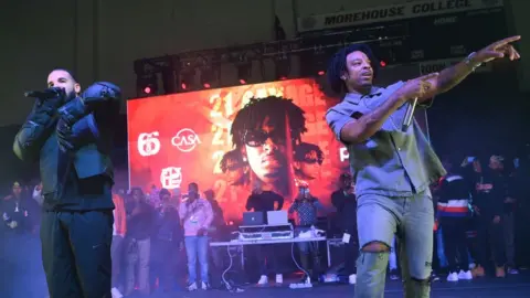 Getty Images Drake and 21 Savage perform in Atlanta Georgia. They're in front of a large, red-hued picture of 21 Savage, showing his face from three different angles, with his name printed repeatedly behind him. Drake is wearing a black bomber-style jacket and thick gloves. His face is mostly obscured as he sings into the microphone. 21 Savage is wearing a short-sleeved shirt and shorts, and pointing at the crowd.