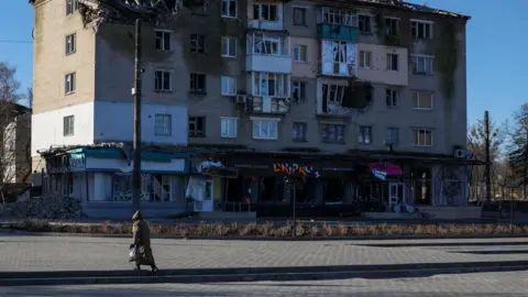 Insulate Ukraine A woman walks across a road towards a housing block damaged by the war in Ukraine. Windows are smashed and masonry is falling away.