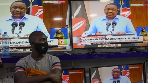 Getty Images A man wearing a face mask stands inside an electronics shop as Kenya's President Uhuru Kenyatta is seen on television screens while giving an address to the nation on the COVID-19 (novel coronavirus) pandemic, and measures that the government maintain and enforce to curb the disease's spread, on July 27, 2020, in Nairobi