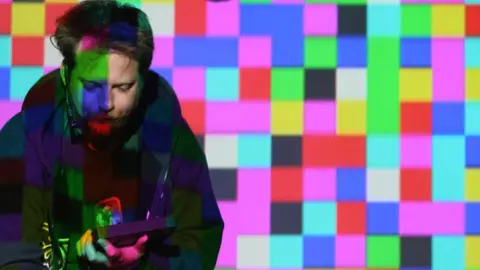 Matthew Applegate A man in front of colourful square lights looks at a handheld device