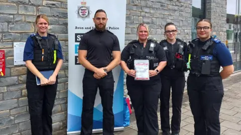 Avon and Somerset Police 5 police officers stood next to a bleed kit which has been installed on the wall behind them. There are 4 women in the image and 1 man. All of them are stood in a line, smiling, looking directly at the camera.