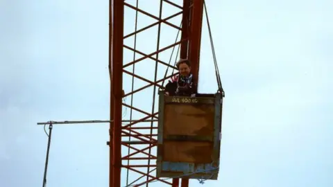 Tony Brien Mr Brien standing with his camera inside a small wooden box suspended high in the air by the crane.