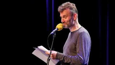 BBC Hugh Dennis standing in front of a microphone