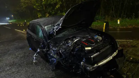 A black car has crashed in a road. The bonnet is up, and the front of the car is crumpled on the divers' side, with bits of metal poking out. The bumper has fallen off.