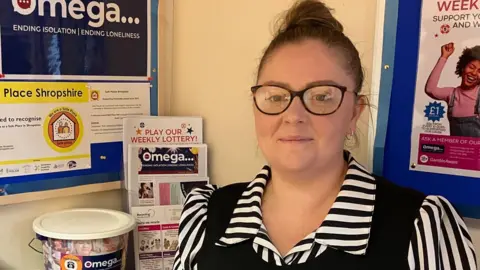 A woman with brown hair and glasses in a black and white striped tops with lots of posters on the wall behind her